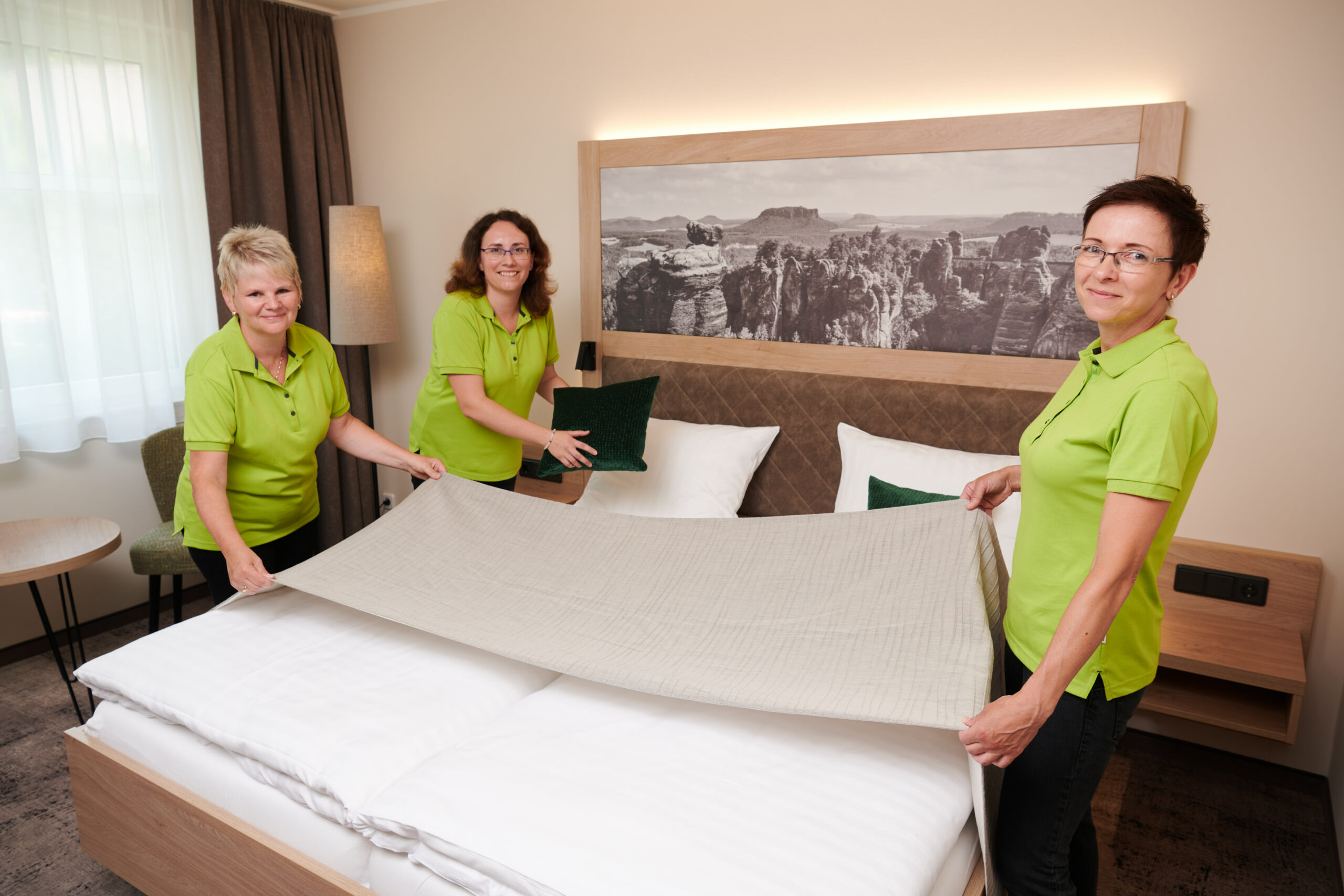 Mitarbeiterinnen des Houskeeping Teams bereiten ein Hotelbett vor und lächeln in die Kamera.