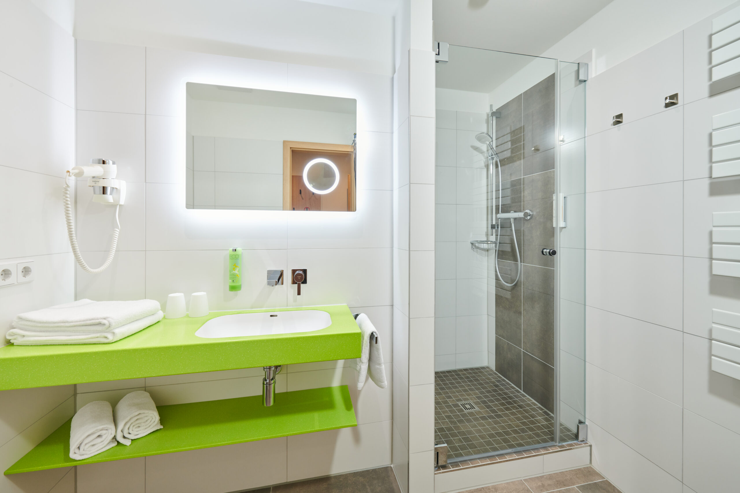 Helles Badezimmer im Parkhotel Neustadt mit weißen Fliesen und einer lebhaften, grünen Ablagefläche. Zur Ausstattung gehören ein weißes Waschbecken, ein großer Spiegel mit integrierter Beleuchtung, eine gläserne Duschkabine mit modernen Armaturen und eine Toilette. Frische Handtücher sind sowohl auf der Ablage als auch an einem Handtuchhalter ordentlich platziert, und ein Haartrockner ist an der Wand befestigt.