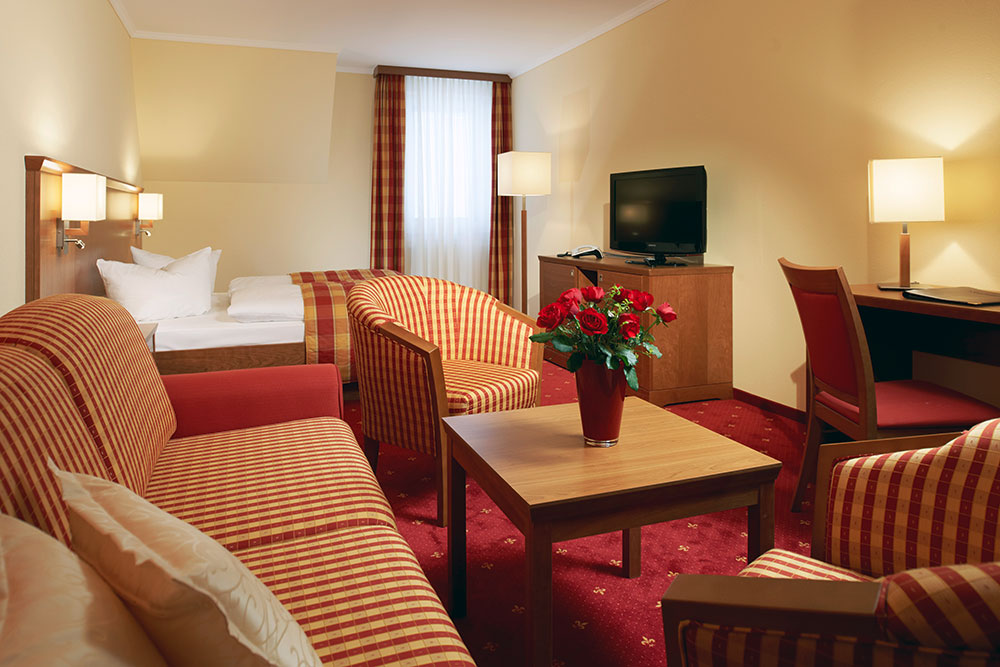 Wohnliche Junior Suite im Parkhotel Neustadt mit einem Doppelbett, gestreifter roter Bettwäsche und einem großen Kopfkissen. Im Vordergrund ein Sitzbereich mit zwei gestreiften Sesseln und einem Sofa um einen Holztisch mit einem Strauß roter Rosen. Ein Schreibtisch mit Lampe und Flachbildfernseher vervollständigen die einladende Einrichtung, ergänzt durch warmes Licht und gemusterte rote Vorhänge, die für eine behagliche Atmosphäre sorgen.